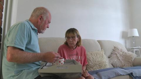 Dave is sat holding a bowl and a tray on his knee as he feeds Carole who is sat on a sofa