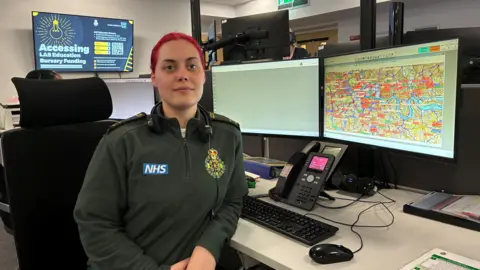 Rose Carbon, an emergency call coordinator, sits in the London Ambulance Service control room. She wears a green uniform and has pink hair. She is sitting at her desk. Her hands are placed on her lap.