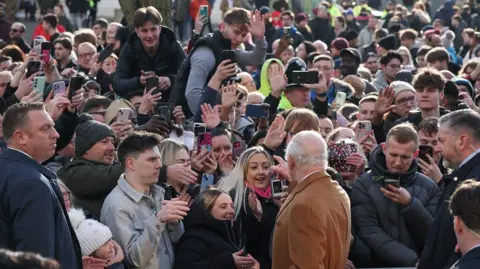 Reuters King Charles standing in front of a crowd of people. He's pointing and hands and phones can be seen waving back at him. 