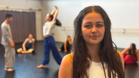Dominic King/BBC A woman with long brown hair and wearing a white top. There are dancers training behind her.