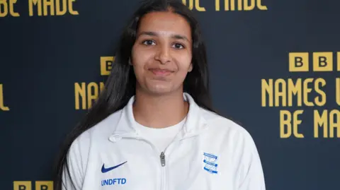 Riya Mannu, a woman wearing a white t-shirt and white sports jacket is stood against a BBC Names Will Be Made step and repeated board which is navy blue with yellow lettering.