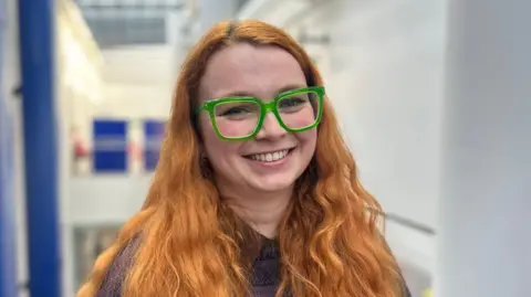 Fiona has ginger hair and is wearing green glasses with a purple coloured top.