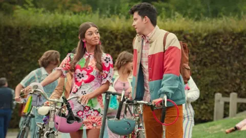 Netflix Mimi Keene as Ruby and Asa Butterfield as Otis in Sex Education. Ruby and Otis are speaking to each other outside as they wheel their bikes. Ruby, a young woman with long light brown hair, wears a short floral dress and large pink earrings. Otis, a young man with short dark hair, wears orange cord trousers, a pink plaid shirt and a blue, cream and red striped puffer jacket.