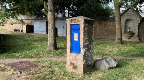 A blue post box set inside a damaged concrete surround with grass, trees, and a stone wall behind it.