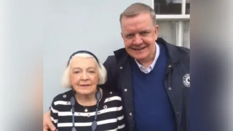 Paul Baker A man with short hair, dark jacket and blue jumper has his arm around an elderly woman who has white hair and a striped black and white top.