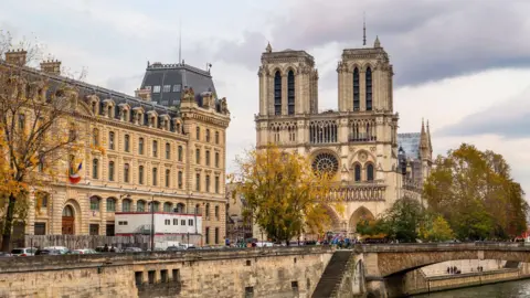 Getty Images The Notre Dame, a large cathedral with two turrets at the front and a circular stained glass window. The photo is taken from the other side of the river, and the water and a bridge are in the bottom right of the photo.