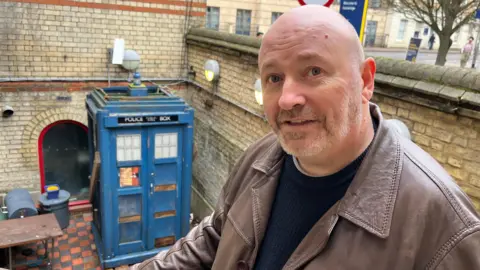 The Reverend Jason de-Vaux is standing on steps above a courtyard where a blue Tardis is placed. He is balding with a short beard and moustache and is wearing a brown leather jacket