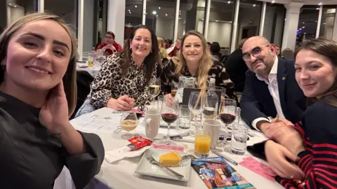 BHA A group of people at a quiz event pose for a selfie as they sit around a round table. On the table in front of them are various glasses, some containing orange juice and red wine and plates of dessert. The table has a white cover on it