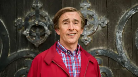 BBC Alan Partridge looking into the camera and smiling. He is wearing a red jacket and a blue and red checked shirt.