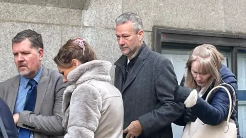 A man with a short brown hair wearing a smart gray coat stands in a line, looking down. A white woman in a blue coat with a cream handbag is behind her, and stands in front of her in front of her in a cute gray coat with a gray suit and a shirt in a gray suit and a shirt. None of them is watching the camera.