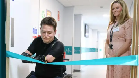 Alex Dixon Photography Alex Lewis cuts a blue ribbon with a pair of scissors. A woman wearing pink and can be seen looking on in the background