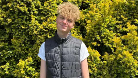 17-year-old Ryan, in a white t-shirt and black gilet, smiles standing in his garden. A large yellow/green bush is behind him.