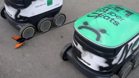 JJ Eteson Two delivery robots with black graffiti. A sad face has been drawn onto one of the robots, which has an Uber Eats logo against a green backdrop.