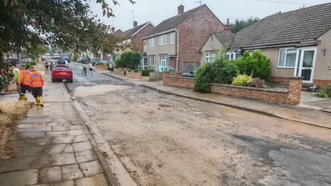 Vincent Clive A road that has mud on it, with rows of houses on either side. Several people are in the street and cars are up the road. 