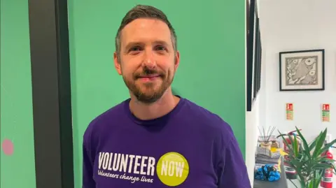 Volunteer now A man, Toby Brusey pictured in an office. He is wearing a purple t-shirt that says 'Volunteer now, volunteers change lives' with a green circle graphic. behind him is a green and white wall with wall art and a plant.