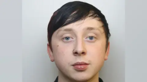 A mug shot of a 22-year-old with limp dyed dark hair, light eyebrows, blue eyes and a black nose ring