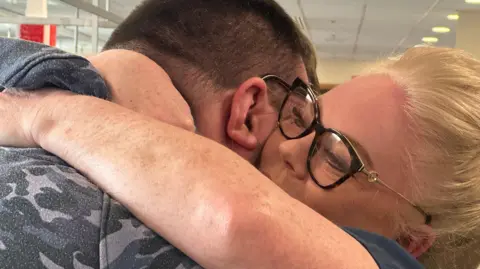 BBC A women with tied back blonde hair wraps her arms around a man wearing a camouflage patterned hoodie. He has short dark hair and you can see the back of his head. She is wearing black rimmed glasses with gold temples and is looking up over his shoulder. 