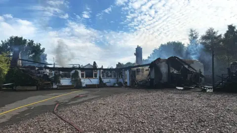 A bungalow that is charred and blackened with parts of its roof collapsed or missing due to a fire. Grey smoke rises from the remains.