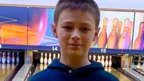 The family is looking at a close-up photo of a boy directly in the camera. He has brown hair and is wearing a blue hoodie. He is in a ten-pin bowling street, which has sets of streets and pins visible behind it.