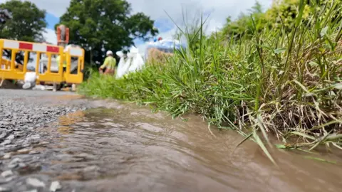 BBC An image of a road in the countryside with water running down the edge of the carriageway. An area of road is fenced off with people in high viz clothing working to fix a leak.