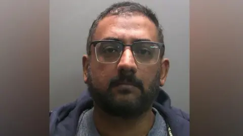 A mugshot of Shamir Chandran. He has short salt and pepper hair and a trimmed beard. He wears glasses with a blue hoodie and grey t-shirt.