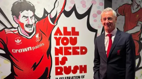 The entrance to the Ian Rush exhibition, decorated with comic-strip illustrations or Ian. Ian is standing in a black suit and red tie, smiling to the camera.
