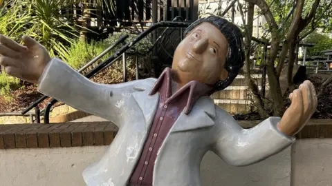 Sculpture of a man with short brown hair wearing a white jacket over a brown shirt with white buttons
