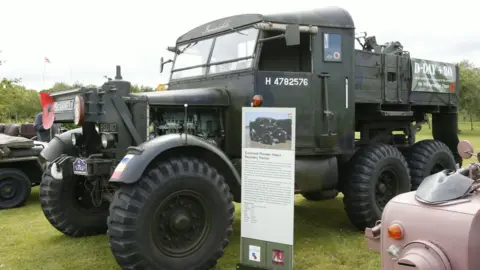 MVT A dark green truck with large black wheels on display in a field. Next to it is a sign with a description and photo of the vehicle. 