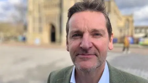 Andrew Sinclair/BBC Pete Waters wearing a green jacket and open necked shirt standing in front of a church in central Norwich 