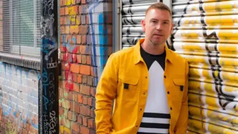 Mark standing outside, wearing a yellow jacket with short hair, standing next to a wall and metal shutters, covered in graffiti on a street 