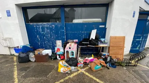 Emma King Household items, such as a doll's house, plastic bags and a desk next to a blue garage door on the wet ground after rain