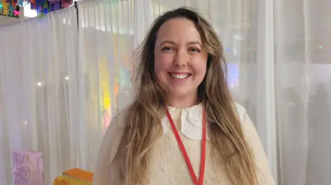 Nikita Dare is standing in front of a white curtain, wearing a white jumper. She has long, blonde hair and is smiling at the camera.