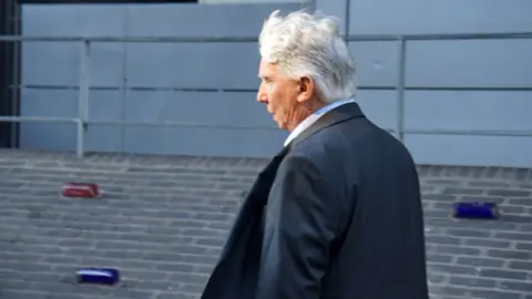 David Geary, a man with gray hair, dressed in a dark suit and white shirt, is walking along a brick pathway. Behind him, a metal railing runs parallel to the path, and the background features a gray wall with horizontal lines, giving the scene a modern, urban appearance.