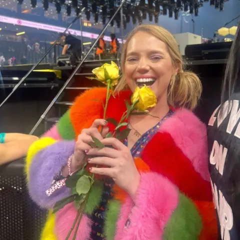 MacKenzie Dunaway Gardner A woman with blonde hair in a brightly-coloured fuzzy cardigan holds two yellow roses in front of a stage at a concert stadium