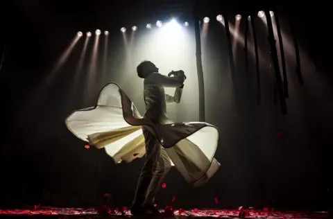 A man dances on stage at The Mount Without in Bristol. He is wearing a white outfit and is illuminated by multiple spotlights