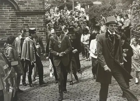 Saltaire World Heritage Education Association A large group of people walking up a cobbled path, led by a man in a top hat and suit, and another in mayoral chains. It's a black and white photo. Others in the group wear ceremonial clothes.
