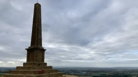 BBC A view from behind the War Memorial, looking out over the landscape below Ham Hill. The memorial itself consists of four steps with a large stone obelisk in the middle. The names of the dead are written on tablets on its side.