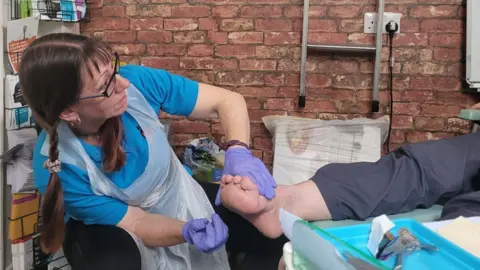 Forgotten Feet Deborah Monk is treating a person in a podiatry session. She is wearing a blue top and plastic apron and has her hair in plaits and is wearing glasses. The person has their leg stretched out on a surface. There is a tray of podiatry equipment next to them.