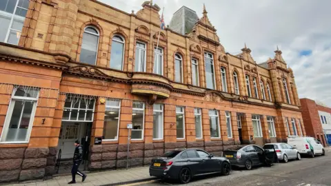 A red brick two-storey building with gables along the front. Cars are parked on the road outside. A man dressed in dark clothing and a beanie hat is walking past the entrance.