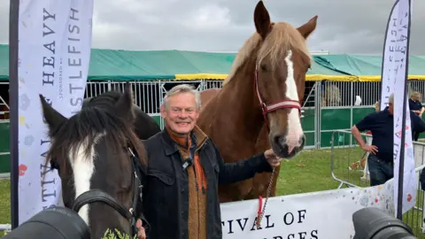 PA Media Martin Clunes is smiling as he holds the bridles of two large horse, one dark brown and one chestnut. 