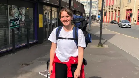 Chloe Davies has her hair tied back and is wearing a white t-shirt and black pants, with a red jacket tied around her waist. She is carrying a large blue and black backpack on her back. She is smiling at the camera as she stands on the sidewalk next to a city road.