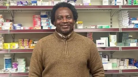 Tunde Olawoye stood behind the counter at his pharmacy wearing a brown fleece jumper. There are shelves of drugs and medicine behind him.