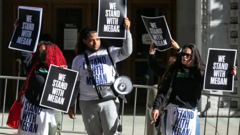 Getty Images Four Megan Thee Stallion supporters stand outside a Los Angeles courthouse. They're each holding a sign with "We Stand With Megan" written in white block capitals on a black background. One, with a megaphone slung over his shoulder, wears a white sweatshirt with the same slogan printed in blue. Three of the protesters stand in front of a metal security railing, while the fourth - who stands behind a fellow protester, is on the other side of it.