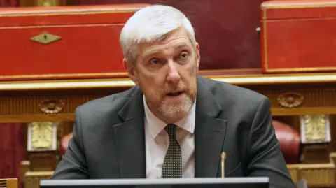 O marcapasso John O'Dowd, com cabelo grisalho curto, vestindo terno cinza e gravata verde, está sentado na câmara do Senado em Stormont.