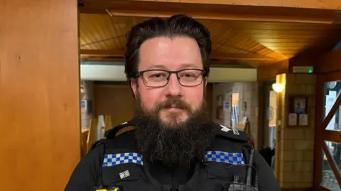 A man with dark hair and a thick dark beard looks at the camera. He's wearing glasses and a police uniform and standing in a church hall.