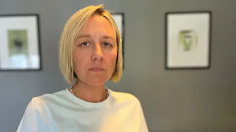 A women is looking directly at the camera. She is sitting on a  chair next to a dining table in a home. She has blonde short hair and is wearing a white T-shirt. She is looking sad. The wall behind her is grey and has three black frames hanging on it. 