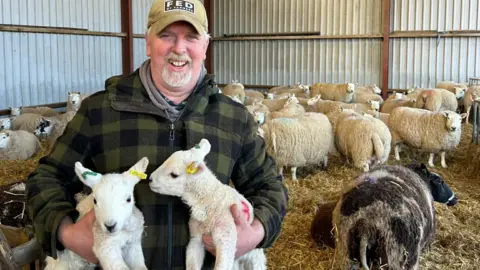 Jim Campbell Sheep farmer Jim Campbell stands in a barn which is filled with sheep which are lying on hay. He is wearing a large tartan green fleece jacket and a beige blonde cap. He is holding two lambs.
