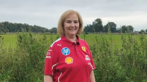 Margaret Peoples is wearing a red ferrari shirt and is standing in a grassy area.
She has long blonde hair.