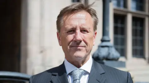 Martin Murray wears a dark grey pinstriped suit jacket, white shirt and grey tie, he is smiling at camera on a sunny day outside a building that is out of focus