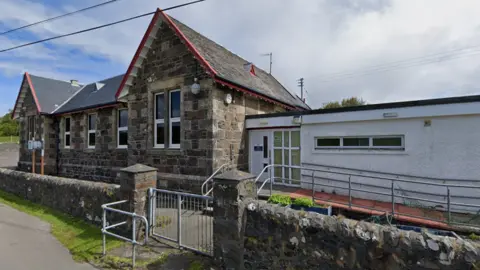 Google A small school building, with a brick wall and gate in front of it